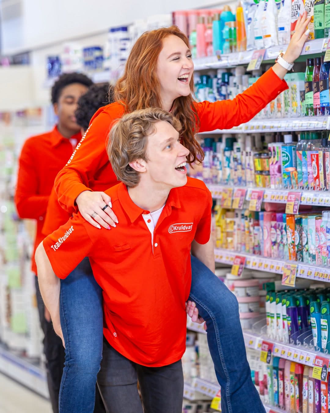 Two Kruidvat employees laughing between the shelves, one on the other’s back